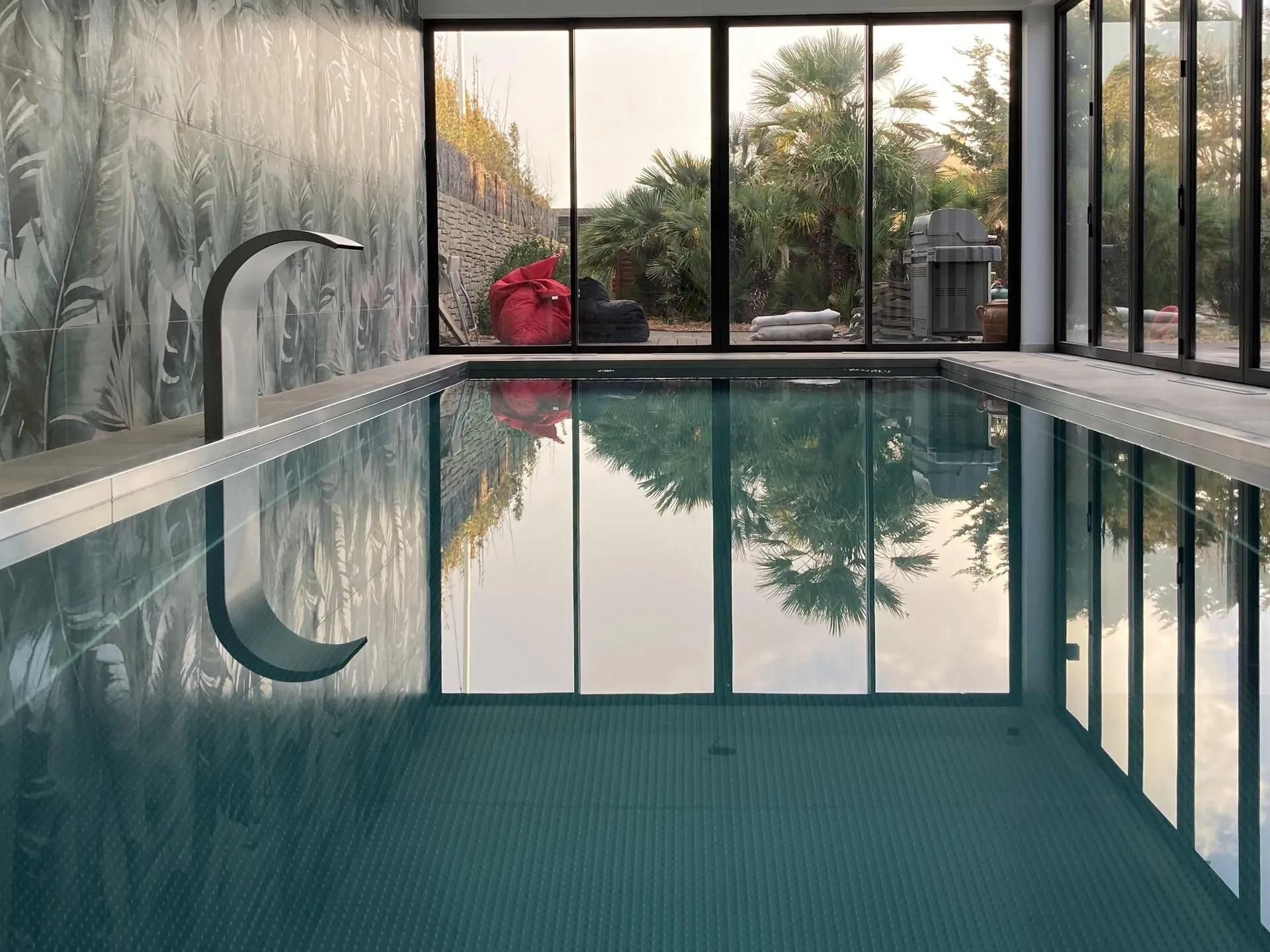 Piscine intérieure à Guérande avec cascade en inox et grandes baies donnant sur des palmiers.